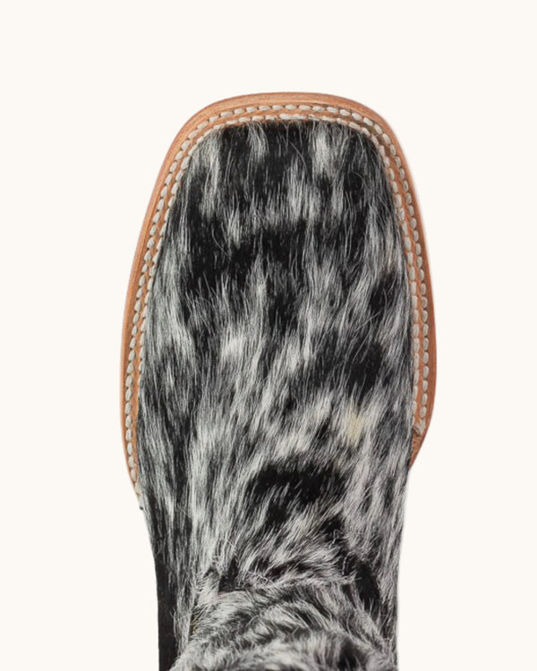 Close-up of the square toe of a western boot featuring a black and white mottled hair-on-hide upper with a tan leather edge and contrast stitching, shown against a white background.